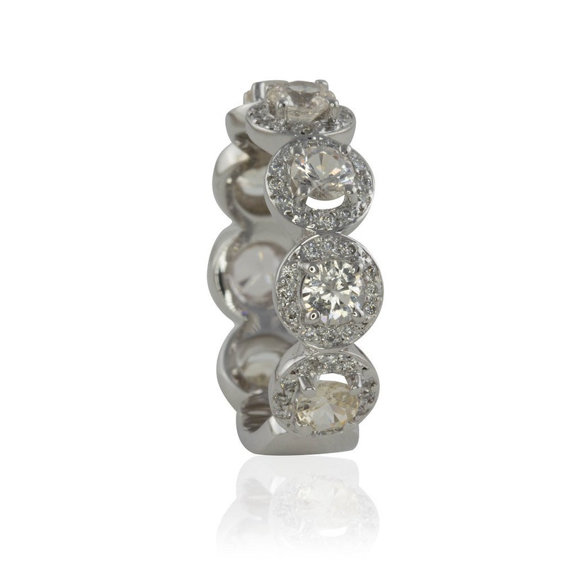 Puede incluir: Un anillo plateado con m&uacute;ltiples piedras preciosas redondas y transparentes. Cada gema est&aacute; rodeada de piedras m&aacute;s peque&ntilde;as y transparentes, creando un efecto de halo. El anillo se encuentra sobre un fondo blanco, con un ligero reflejo.