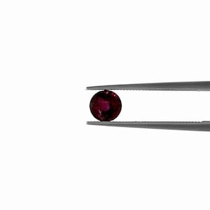May include: A round, dark red gemstone held by silver tweezers against a white background. The gemstone has a deep, rich color with subtle variations in tone, suggesting internal facets and light play. The tweezers provide a clean, professional presentation.