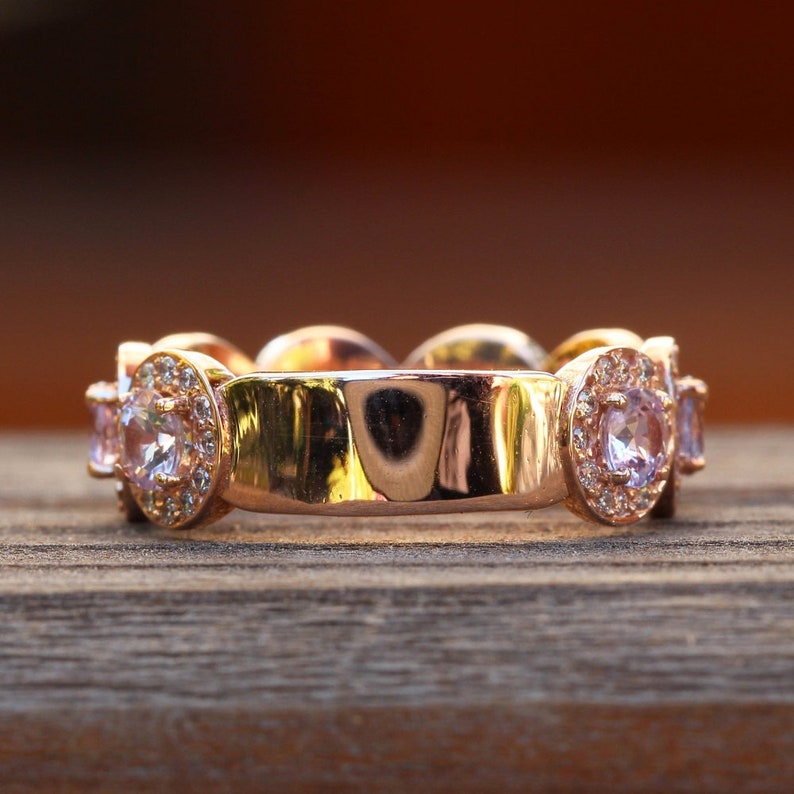 Puede incluir: Un anillo de oro rosa con una serie de piedras preciosas de color p&uacute;rpura claro y peque&ntilde;as piedras transparentes. El anillo est&aacute; colocado sobre una superficie de madera con un fondo marr&oacute;n borroso. El dise&ntilde;o del anillo presenta un patr&oacute;n repetitivo de piedras preciosas.