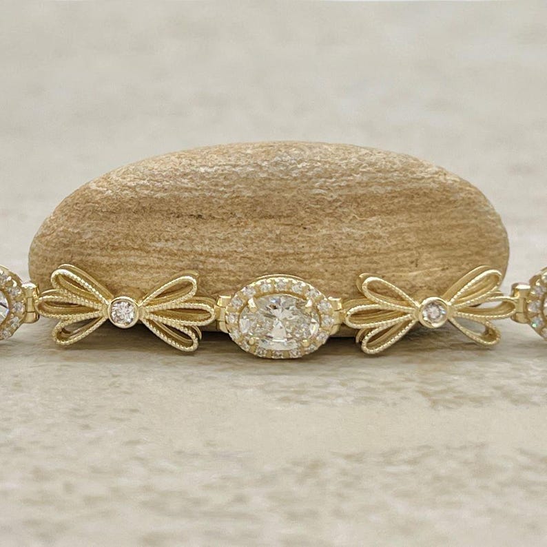 May include: A gold bracelet with a central oval-shaped diamond surrounded by smaller diamonds. The bracelet is decorated with bow-shaped accents on either side of the diamond.