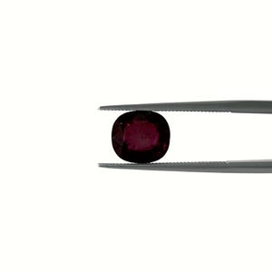 Peut inclure: Une pierre précieuse ovale rouge foncé, tenue par une pince argentée. La pierre a une surface polie et une couleur riche, avec un centre légèrement plus clair. La pince est positionnée sur un fond blanc uni.