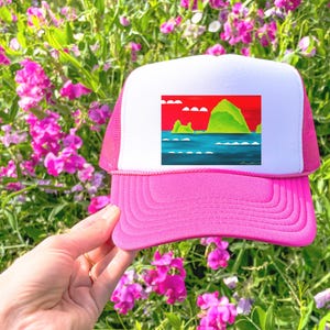 May include: A pink and white trucker hat with a colorful illustration of a green island with a red sky and blue water. The hat has a mesh back.
