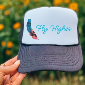 May include: A trucker hat with a white front panel displaying a colourful feather graphic and the words "Fly Higher" in turquoise script. The brim and mesh back are dark grey, with a black rope detail. The hat is held up by a hand.