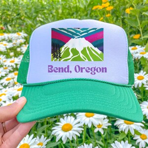 Op de afbeelding: Groene en witte truckerpet met een afbeelding van een berg en bomen. De tekst "Bend, Oregon" staat op de voorkant van de pet. De pet heeft een groene klep en een mesh achterkant.