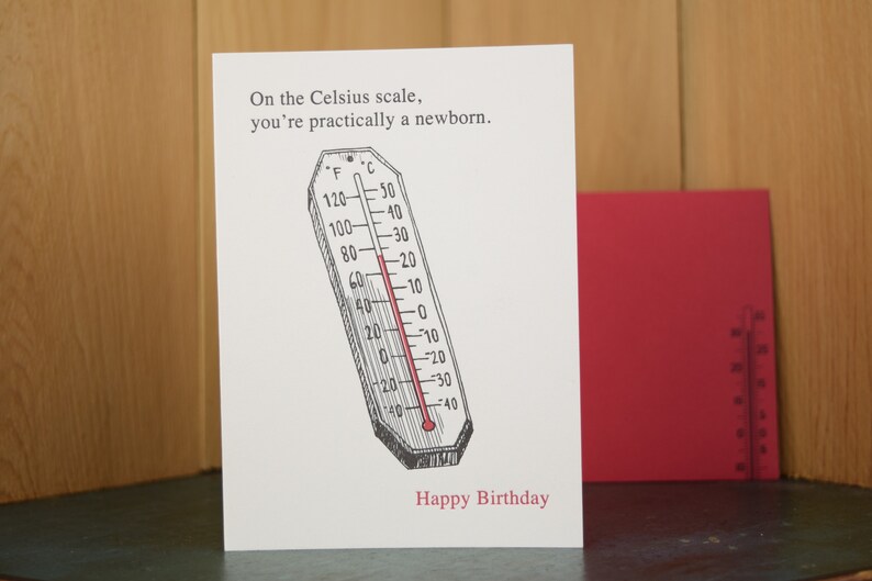 May include: A white card with a black and white illustration of a thermometer with a red line indicating a temperature. The text on the card reads "On the Celsius scale, you're practically a newborn." and "Happy Birthday".