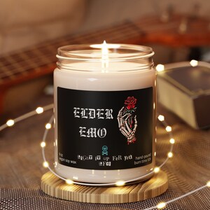 May include: A black candle with a skull and rose graphic and the text "ELDER EMO" on the label. The candle is in a clear glass jar and is sitting on a wooden coaster.