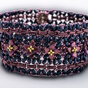 Può includere: Un braccialetto di perline con uno schema di colori viola e nero. Il braccialetto presenta un motivo ripetitivo di piccoli fiori con centri dorati.