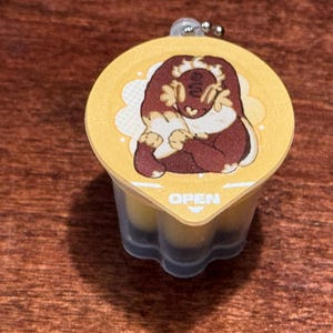 May include: A yellow container with a cartoon illustration of a brown creature with a white outline. The container has the word "OPEN" printed on it. The container is attached to a silver keychain.
