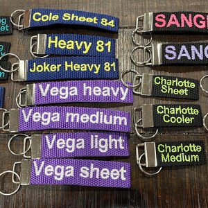 May include: A collection of 18 colorful keychains with various text, including "Cole Sheet 84", "Heavy 81", "Joker Heavy 81", "Vega heavy", "Vega medium", "Vega light", "Vega sheet", "Sangria", "Charlotte Sheet", "Charlotte Cooler", "Charlotte Medium", "Shadow Sheet", "Shadow Cooler", "Shadow Medium", "Ming Sheet", "Ming Heavy", "Ming Medium", and "Snow Sheet", "Snow Heavy", "Snow Medium".