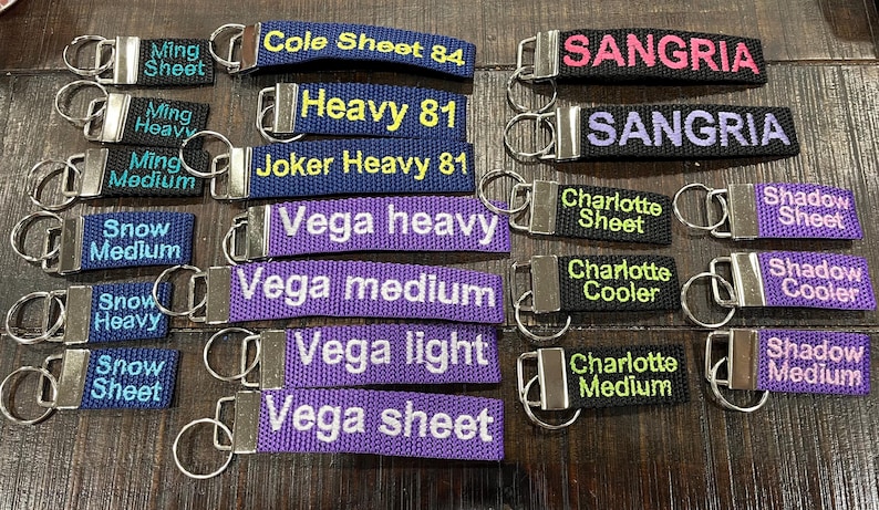 May include: A collection of 18 colorful keychains with various text, including "Cole Sheet 84", "Heavy 81", "Joker Heavy 81", "Vega heavy", "Vega medium", "Vega light", "Vega sheet", "Sangria", "Charlotte Sheet", "Charlotte Cooler", "Charlotte Medium", "Shadow Sheet", "Shadow Cooler", "Shadow Medium", "Ming Sheet", "Ming Heavy", "Ming Medium", and "Snow Sheet", "Snow Heavy", "Snow Medium".
