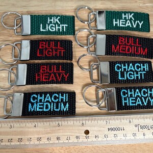 May include: Ten keychains with black and green fabric straps and silver key rings. The straps have white, red, and blue embroidered text that reads "HK Light", "HK Heavy", "Bull Light", "Bull Medium", "Bull Heavy", "Chachi Light", "Chachi Medium", and "Chachi Heavy".