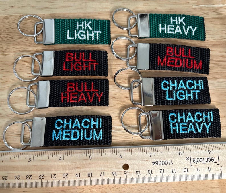 May include: Ten keychains with black and green fabric straps and silver key rings. The straps have white, red, and blue embroidered text that reads "HK Light", "HK Heavy", "Bull Light", "Bull Medium", "Bull Heavy", "Chachi Light", "Chachi Medium", and "Chachi Heavy".