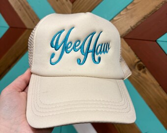 May include: A beige trucker hat with a mesh back and a curved brim. The hat features the embroidered text "Yee Haw" in a teal script across the front. The hat is held up against a wooden background with teal, brown, and white accents.