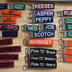 May include: A collection of colorful keychains with embroidered names and phrases. The keychains are made of woven fabric and have silver metal key rings. Some of the names include "Aspen", "Ruby", "Ned Joe", "Scotch", "Whiskey", "Pixie 72" and "Cody".