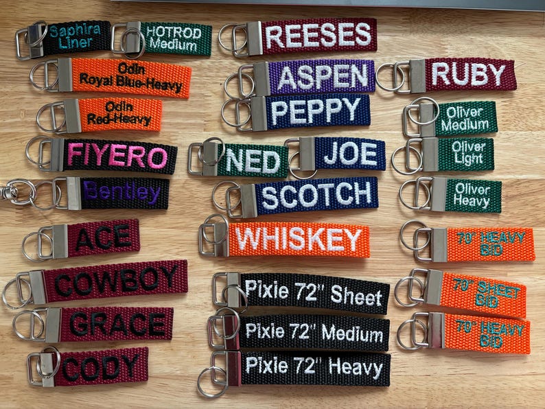 May include: A collection of colorful keychains with embroidered names and phrases. The keychains are made of woven fabric and have silver metal key rings. Some of the names include "Aspen", "Ruby", "Ned Joe", "Scotch", "Whiskey", "Pixie 72" and "Cody".