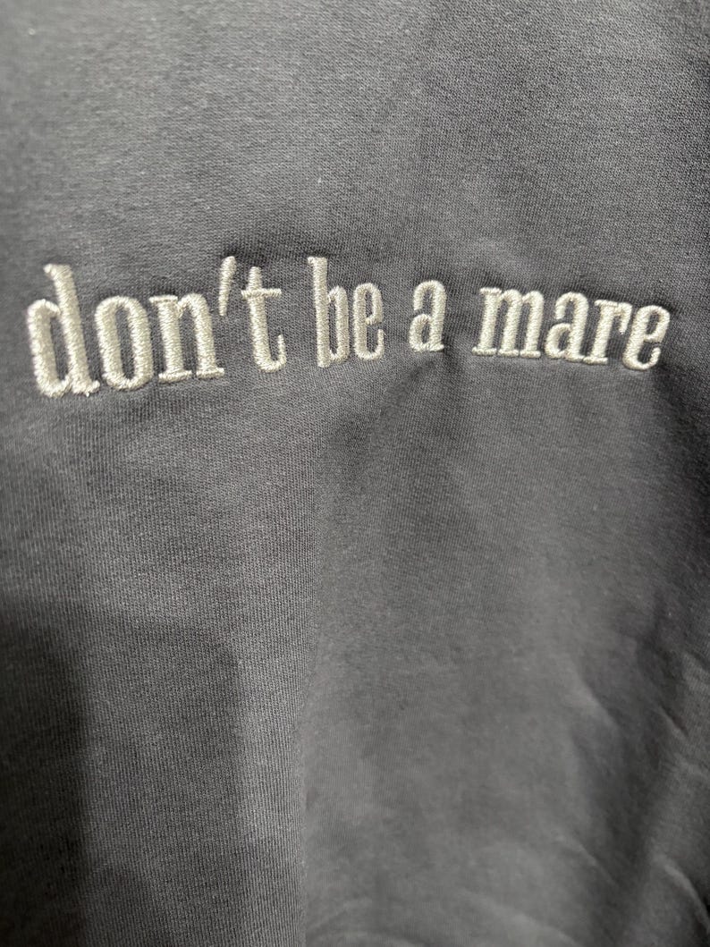Puede incluir: Una sudadera gris oscuro con el texto "don't be a mare" bordado en blanco.