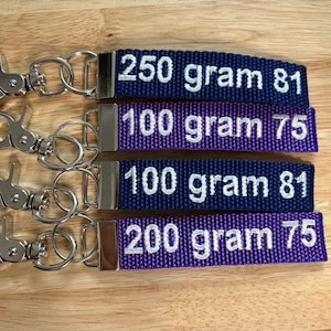 May include: Four keychains with different weight labels. The keychains are purple and blue with white text. The labels read "250 gram 81", "100 gram 75", "100 gram 81", and "200 gram 75".