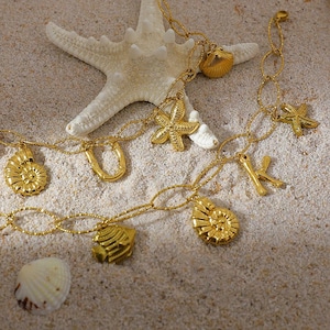 Könnte beinhalten: Eine goldene Kettenkette mit mehreren Anhängern, darunter Muscheln, Seesterne und ein Bambusdesign. Die Kette wird auf einer sandigen Oberfläche mit einem weißen Seestern gezeigt.