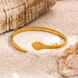Puede incluir: Un brazalete de puño de serpiente de color dorado. El brazalete presenta un diseño de cabeza de serpiente texturizada y un cuerpo texturizado. El brazalete está abierto en la parte inferior y descansa sobre una superficie arenosa con una concha y madera flotante.