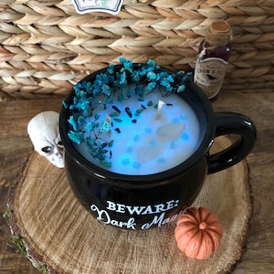 May include: Black cauldron-shaped mug candle with a blue wax pool, teal dried flowers, and a small white crystal. The mug has the text "BEWARE: Dark Magic" on the side. A small orange pumpkin sits on a wooden surface.