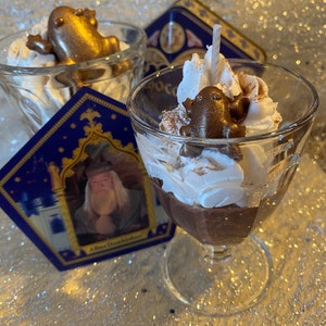 May include: A glass mug filled with hot chocolate topped with whipped cream and a chocolate frog figurine. The mug is sitting on a table with a decorative coaster featuring a wizard and the text "Albus Dumbledore".