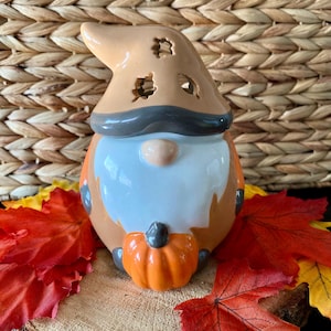 May include: A ceramic gnome figurine with a brown hat, gray beard, and a white face. The gnome is holding a small orange pumpkin. The gnome is sitting on a wooden slice with red and orange fall leaves.