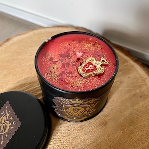 Puede incluir: Una vela roja con purpurina dorada en un recipiente de hojalata negro. La tapa del recipiente tiene un escudo de león dorado con una corona en la parte superior. La vela está decorada con un amuleto dorado en forma de escudo de león.