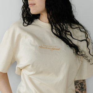 Pode incluir: Uma mulher vestindo uma camiseta creme com o texto "We were born for all the suburban legends" em laranja. Ela está usando jeans preto e tem cabelo comprido e escuro.