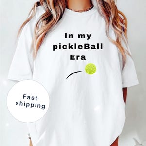 Può includere: T-shirt bianca con la scritta "In my pickleBall Era" in nero. Una palla da pickleball gialla con dettagli verdi è raffigurata accanto a una linea nera curva. La maglietta è in stile casual.