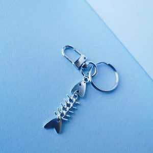 Może przedstawiać: Srebrny brelok do kluczy w kształcie szkieletu ryby z karabińczykiem i dzielonym kółkiem. Projekt szkieletu ryby ma ogon, głowę i kręgosłup. Brelok znajduje się na jasnoniebieskim tle.