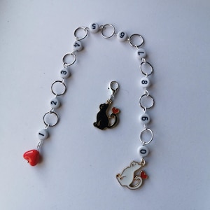 May include: A silver chain bracelet with white beads numbered 0 through 9. A black cat charm with a red heart is attached to the chain. A white cat charm with a red heart is attached to the chain.