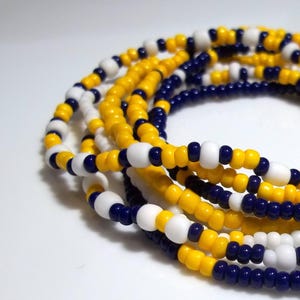 May include: Strands of beaded jewelry in a repeating pattern of yellow, white, and navy blue. The beads are small and round, creating a textured surface. The jewelry is likely worn around the waist or as a necklace.