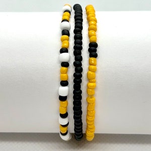 May include: Three beaded bracelets in black, white, and yellow. The bracelets are stacked on top of each other.