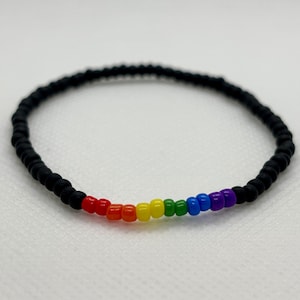 May include: A beaded bracelet featuring a rainbow of colors, transitioning from red to purple, set against a backdrop of black beads. The bracelet is made of small, round beads and is designed to be worn around the wrist.