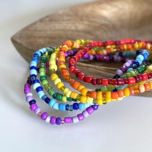 May include: A collection of colorful beaded bracelets in a rainbow of hues, including red, orange, yellow, green, blue, and purple. The bracelets are displayed on a wooden surface, showcasing the vibrant colors and intricate beadwork.