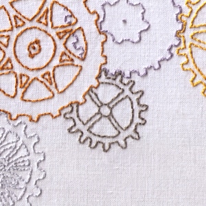 Puede incluir: Diseño bordado de engranajes entrelazados en tonos naranja, gris y morado sobre un fondo de tela blanco.