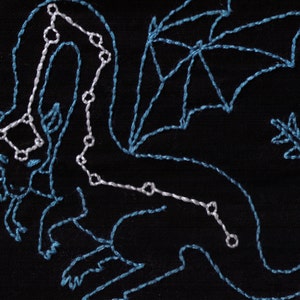 May include: Embroidered design of a blue dragon on a black fabric background. The dragon is outlined in blue thread and has a white constellation pattern on its body.