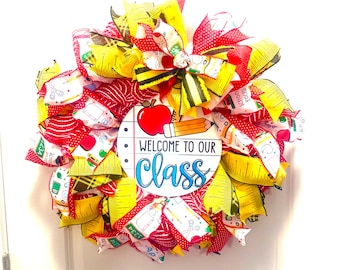 May include: A colorful school-themed wreath with red, yellow, and white ribbons. The wreath features a central circular sign with an apple, pencil, and the words "Welcome to Our Class" in blue.