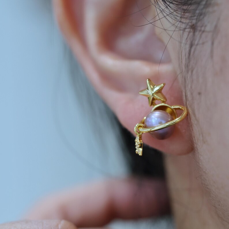May include: Gold-colored celestial-themed earring featuring a star and a planet design. The planet is a light purple pearl. The earring is attached to the earlobe.