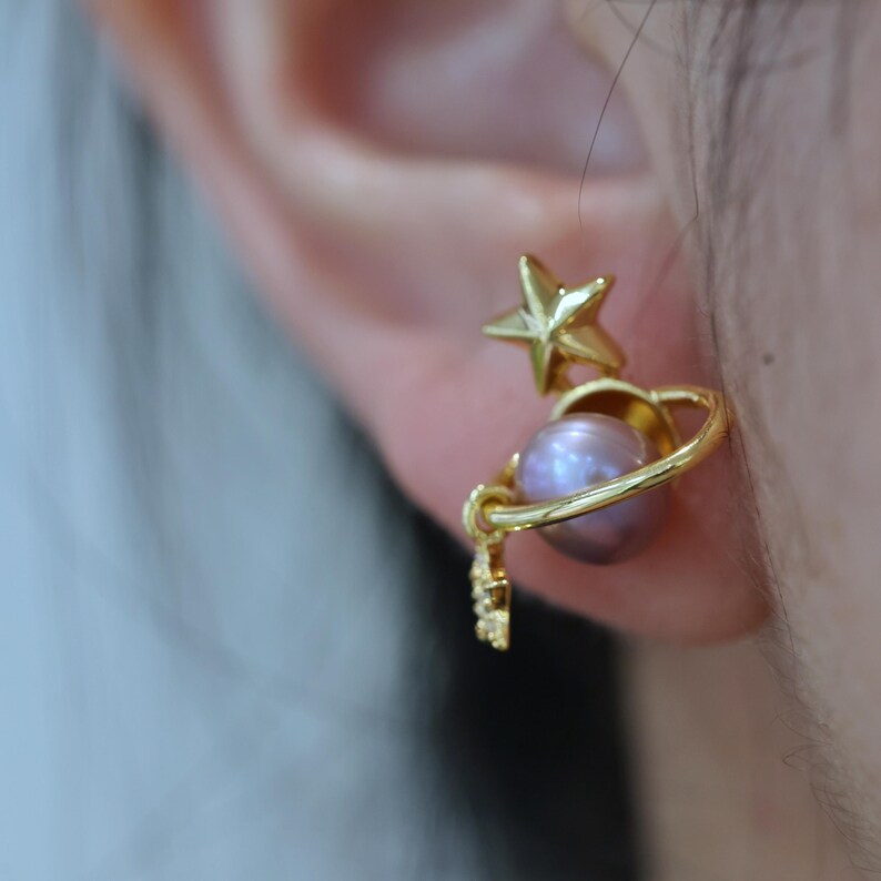 May include: Gold-toned celestial earrings featuring a star and a planet design. The earrings have a star stud and a planet with a lavender-colored pearl. The planet is encircled by a gold ring, with a small decorative element.