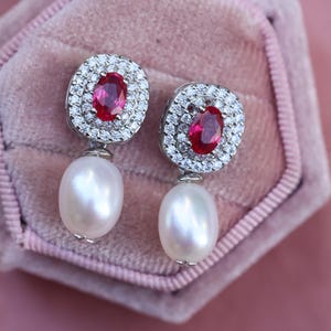 Puede incluir: Un par de pendientes elegantes con piedras rojas ovaladas rodeadas por un halo de pequeñas piedras brillantes. Cada pendiente cuelga una perla grande, brillante, en forma de lágrima. Sobre un fondo rosa suave.