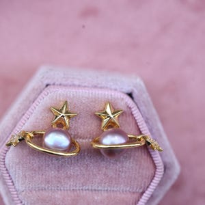 May include: A pair of gold-colored earrings with a celestial theme. Each earring features a star-shaped top, a light purple pearl, and a gold ring encircling the pearl. The earrings are set against a pink velvet background.