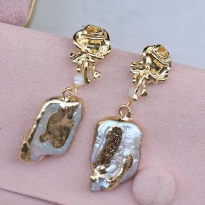 May include: Gold earrings featuring a rose design and a large, irregular pearl drop. The pearls are white with a slight iridescent sheen.