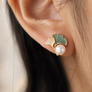 Peut inclure: Une boucle d'oreille à tige dorée présentant deux formes de feuilles de ginkgo, l'une en émail crème et l'autre en émail vert sauge, agrémentée d'une perle blanche ronde. La boucle d'oreille possède une tige dorée reliant les feuilles et la perle.