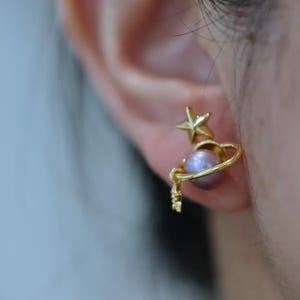 May include: Gold-colored celestial earrings. One earring features a star and a planet design with a lavender pearl. The other earring is out of focus. The earrings are worn on a person's ear.