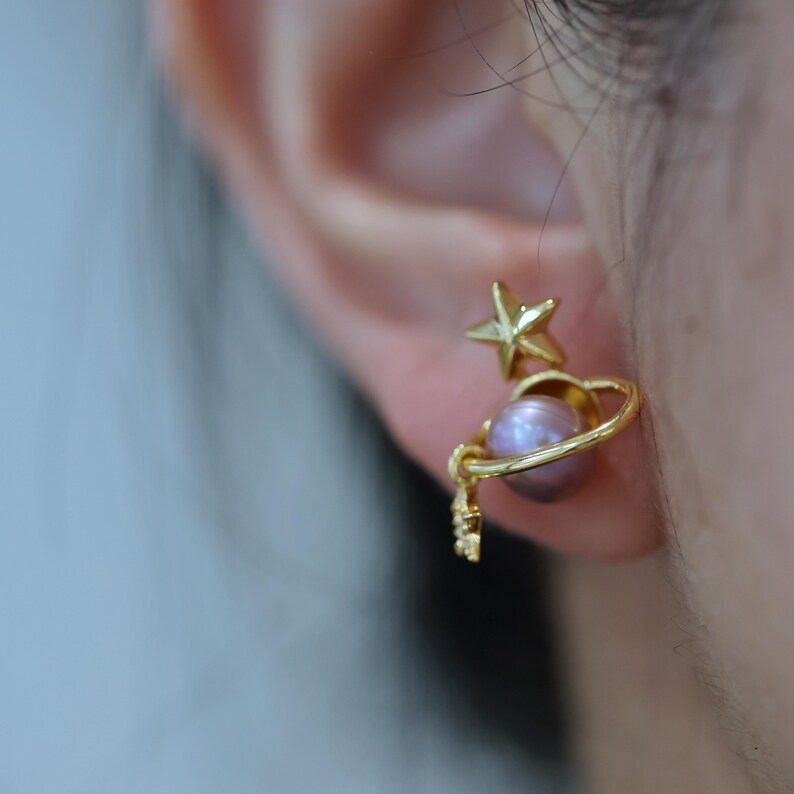 May include: Gold-colored celestial earrings. One earring features a star and a planet design with a lavender pearl. The other earring is out of focus. The earrings are worn on a person's ear.
