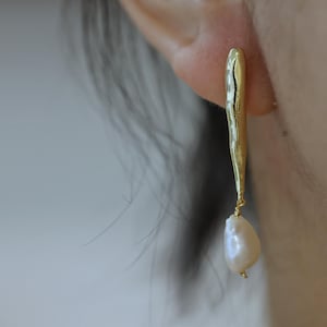 Puede incluir: Pendientes de aro dorados con una barra texturizada y una perla blanca.