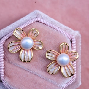 Puede incluir: Un par de pendientes de oro en forma de flor con una perla en el centro de cada flor. Las flores son rosas y blancas con un acabado brillante.