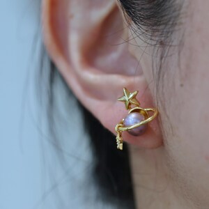 May include: Gold-colored stud earrings featuring a celestial design. Each earring has a star, a planet-like sphere with a lavender hue, and a small dangling charm. The earrings are worn on an ear.