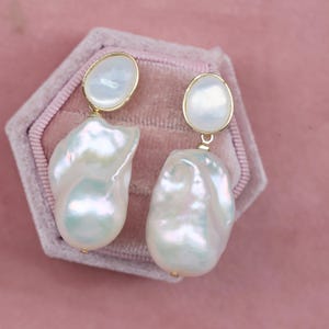 May include: A pair of drop earrings featuring a smooth, round white pearl stud connected to a large, irregular baroque pearl pendant. The pearls exhibit a lustrous, iridescent finish with subtle pink and blue undertones, secured by gold-toned metal settings.